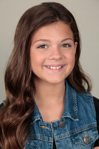 Young actress with a joyful smile, long wavy brown hair, wearing a denim vest in a studio headshot from Ken Weingart's Los Angeles headshots series.