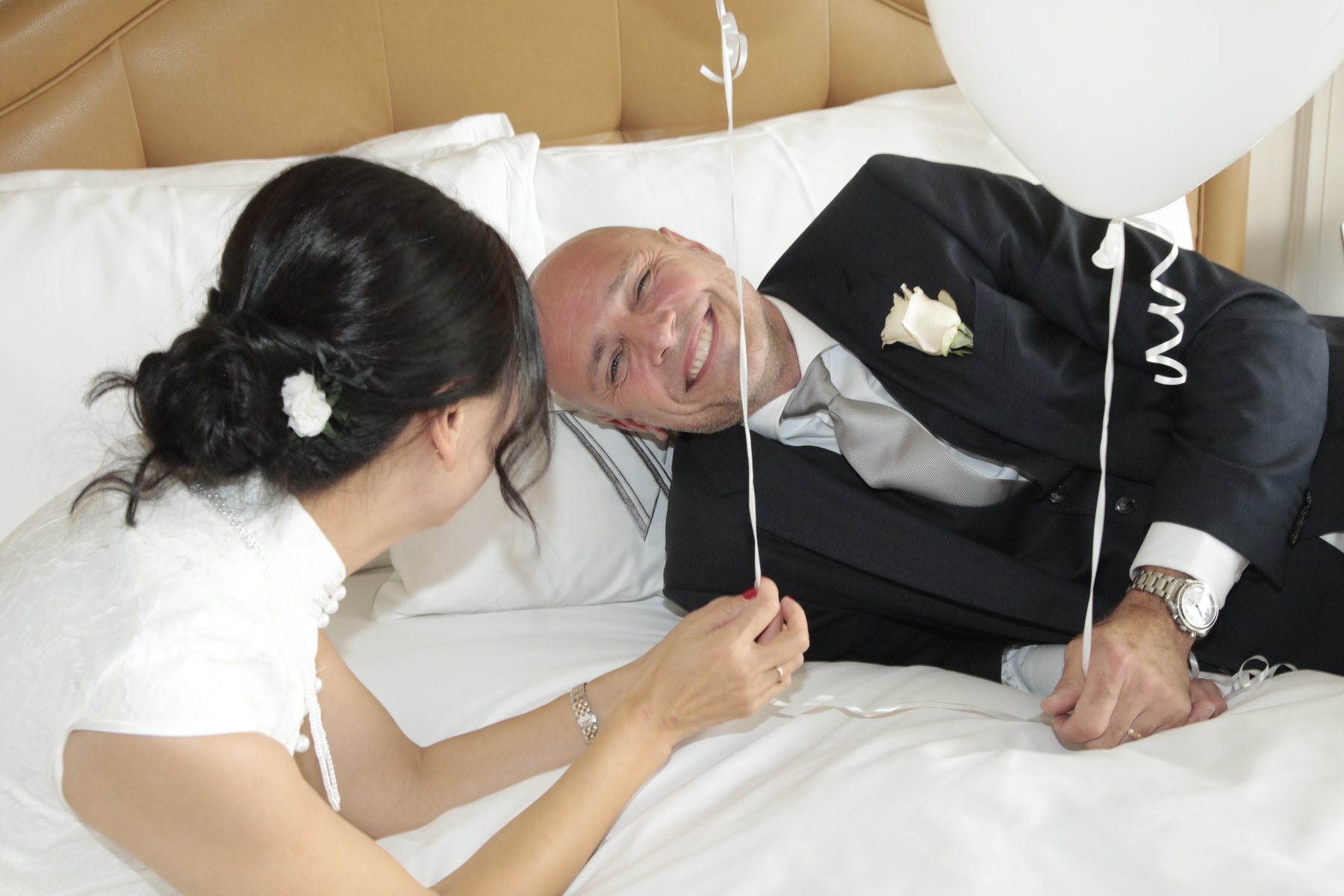 Playful candid wedding portrait of bride and groom with white balloon on hotel bed